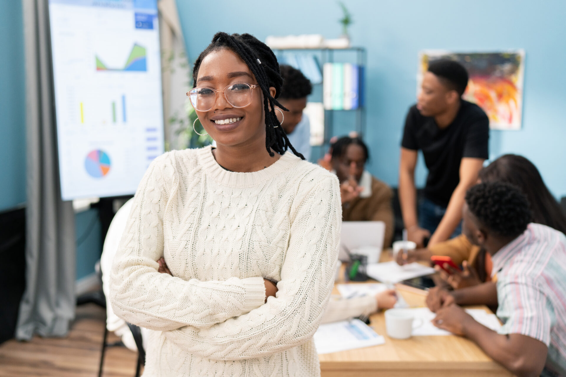 Portrait of a smiling dark-skinned girl wearing a warm sweater with her arms crossed over chest, woman wears glasses and gold earrings, her hair is braided, a business meeting in office in background image of a team of tech professionals