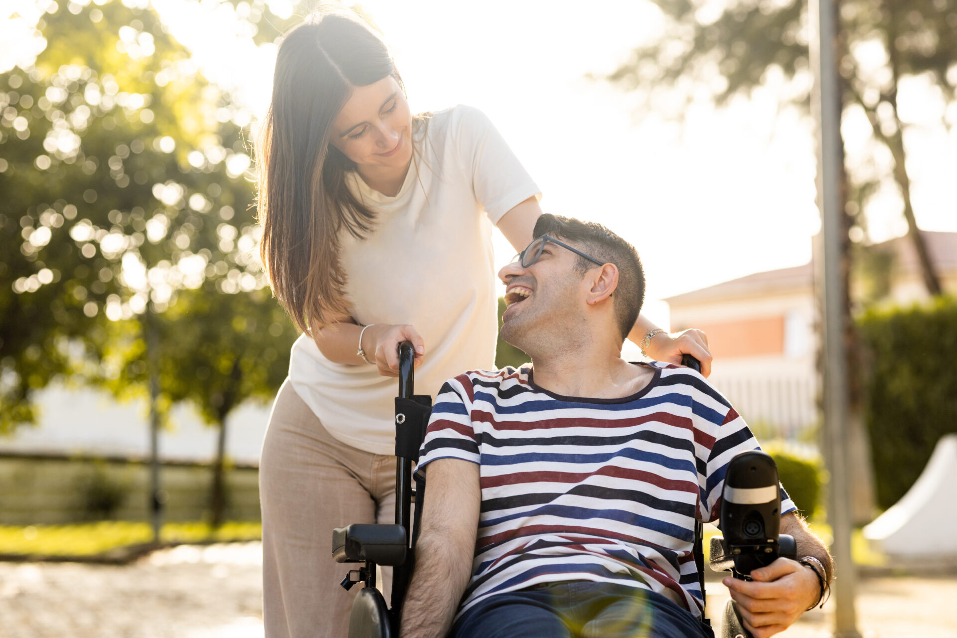 An adult man with a disability sits in an automatic wheelchair with a young woman in a park at sunset.The woman and the man look at each other happily. Concept of support for disabled people. Join DiverseNote Mobility